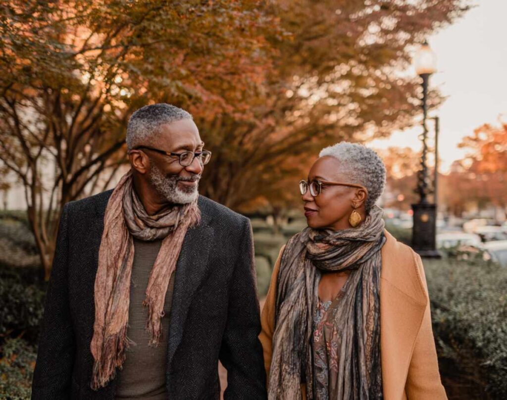 A stylish Black couple aged 55 to 60 walking hand in hand through an upscale city park at golden hour.